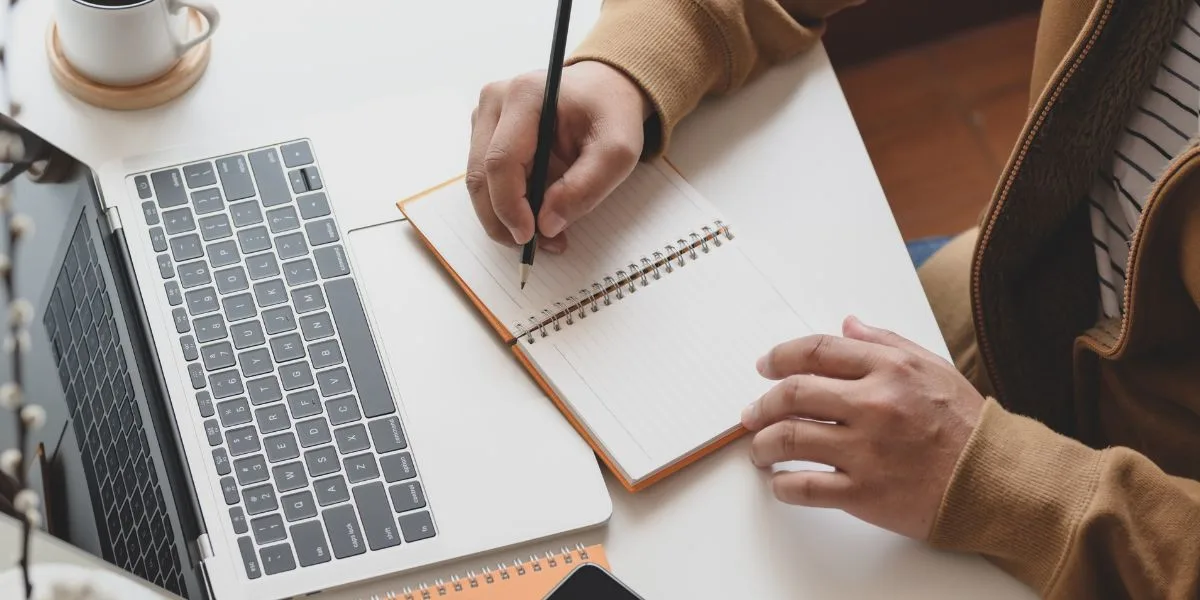 Close up of a desk, laptop with a webinar open, and the hands of man taking notes as he watches.