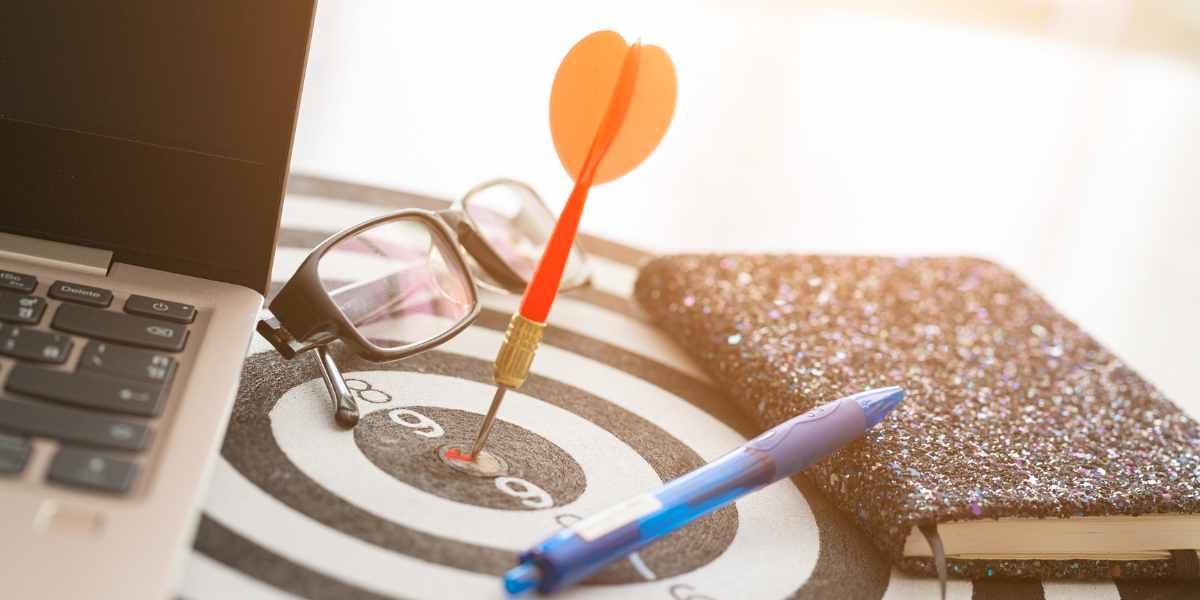 A dart sits at the bullseye of a target on a desk, next to a laptop, glasses, pen, and notebook.