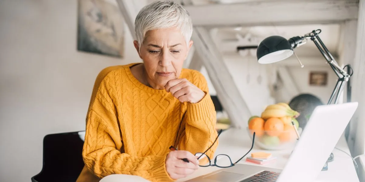 Mature woman in yellow sits at her desk, glasses in hand, in thought about her Medicare choices.