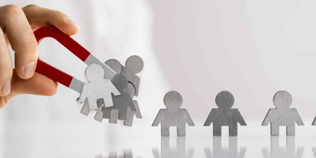 Hand appears on the left side of the image holding a magnet. Small magnetic people are lined up and being pulled up.